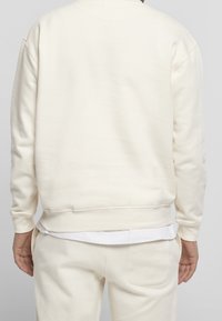 Felpa color crema chiaro con una texture morbida, polsini e vita a costine. Presenta una vestibilità rilassata e un design minimale.