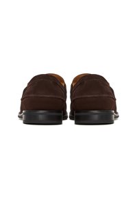 Paire de mocassins en daim marron foncé vus de dos, avec des talons empilés noirs et des coutures visibles sur fond blanc.