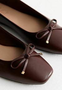 Un paio di ballerine in pelle marrone scuro con punte quadrate e fiocchi, dotate di piccole punte metalliche dorate, su una superficie chiara.