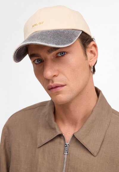 Man med blå ögon som bär en beige och grå keps med texten "NN.07", brun zip-tröja och silverringörhänge som tittar mot kameran.