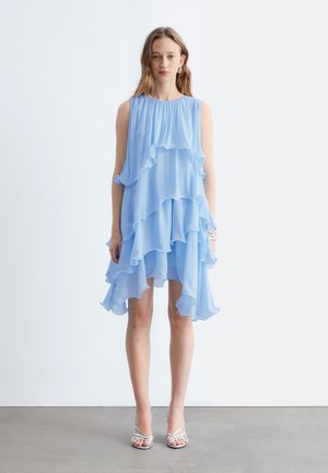 Młoda kobieta stojąca, ubranaw bezrękawną, jasnoniebieską, warstwową sukienkę z falbankami i srebrne sandały na obcasie z paskami na tle jednolitego białego tła.