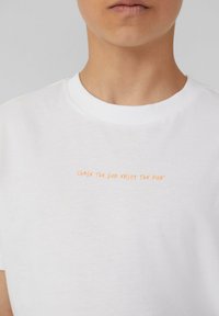 Witte katoenen T-shirt met een ronde hals, met daarop oranje tekst die luidt: "CHASE THE SUN ENJOY THE FUN" in een casual lettertype.