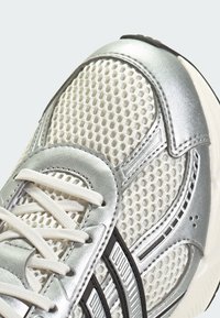 Detailný záber strieborného a bieleho športového topánky s sieťovinou, bielymi šnúrkami a čiernym pruhovaným dizajnom na boku.