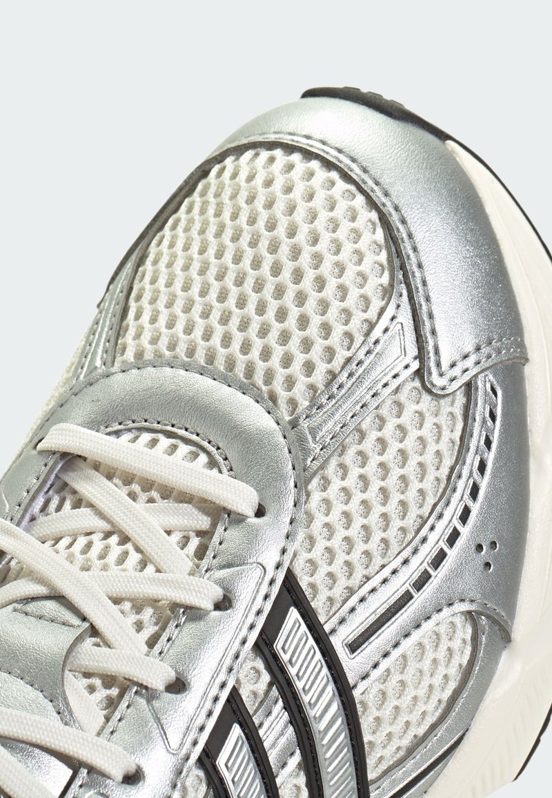 Close-up van een zilveren en witte sportschoen met gaasstof, witte veters en een zwart gestreept ontwerp aan de zijkant.