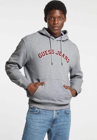 Felpa grigia con "GUESS JEANS" ricamato in rosso, cappuccio con cordini regolabili, tasca frontale e polsini a costine. Abbinata a jeans azzurri.