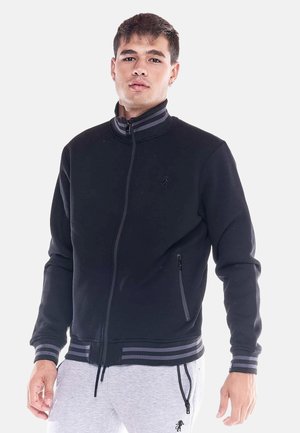 Joven con una chaqueta negra con cremallera, puños y cuello a rayas, combinada con pantalones gris claro con bolsillos con cremallera.