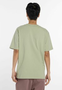 T-shirt en coton vert clair, à manches courtes, avec un col rond et une coupe décontractée. Les détails notables incluent une texture naturelle et des coutures minimales.