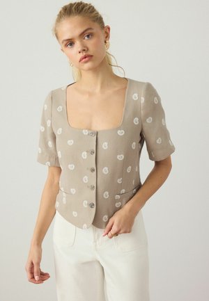 Fiatal, szőke hajú nő bézs színű, rövid ujjú gombos blúzt, fehér paisley mintával és fehér nadrágot visel, sima háttér előtt áll.