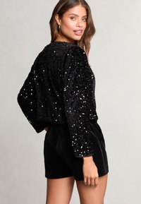 Schwarzes, paillettenbesetztes Crop-Top mit langen Ärmeln, kombiniert mit schwarzen Samt-Shorts. Die Textur ist glänzend und reflektierend, was das visuelle Interesse steigert.