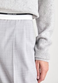Graue karierte Hose mit einem weißen elastischen Bund, auf dem das Logo "MarcCain Sports" zu sehen ist. Der Pullover ist hellgrau mit gerippten Bündchen.