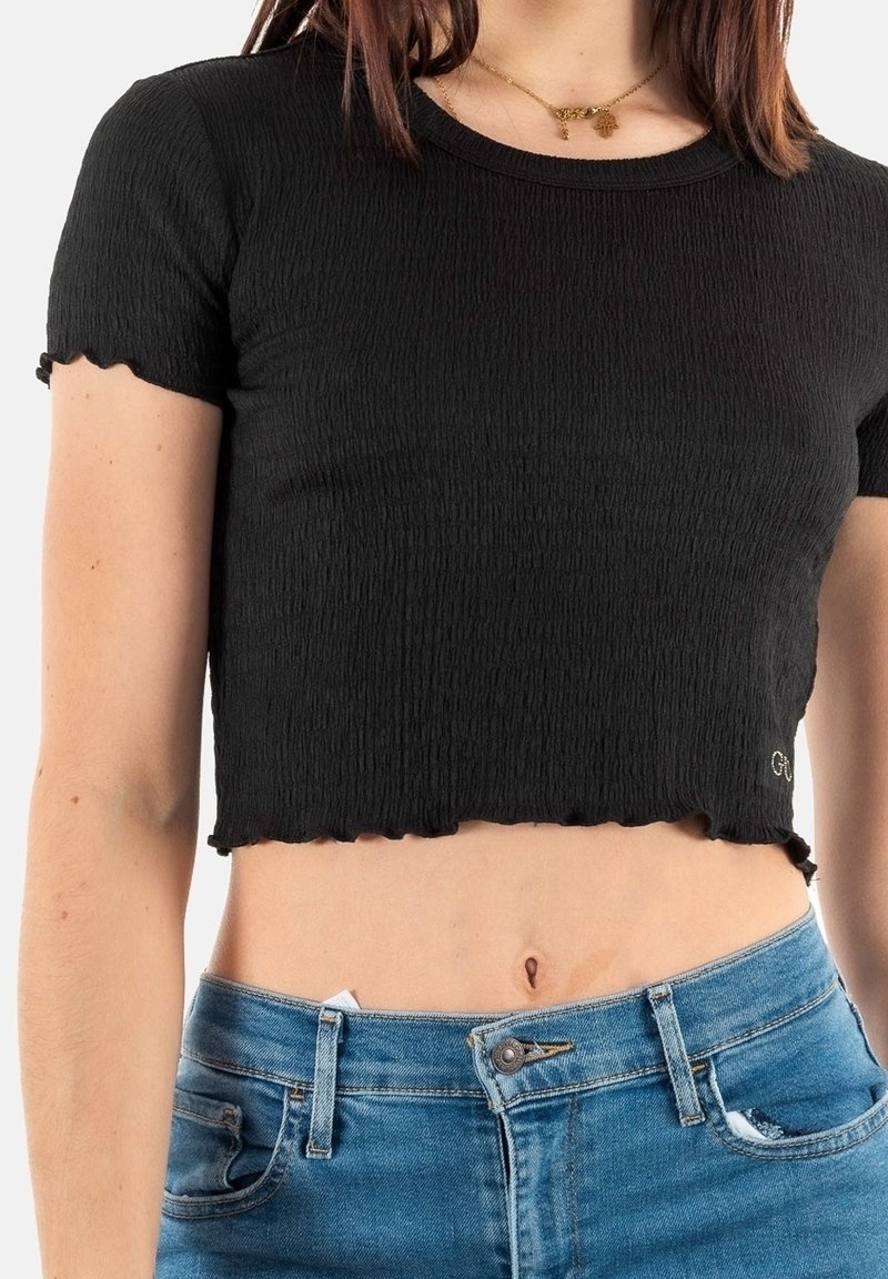 T-shirt noir court en tissu texturé avec des bords festonnés. Avec un col rond et des manches courtes. Associé à un jean en denim bleu.