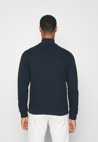 Marinblå sweatshirt med hög krage, ribbade ärmslut och fåll. Mjuk textur, enkelt design. Utsikt från baksidan, bärs över ljusa byxor.