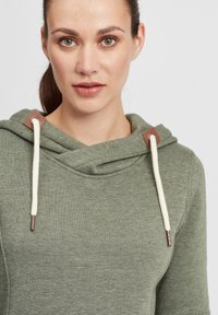 Grön huva-pullover tillverkad av mjukt tyg, med vita dragsko och bruna accenter vid halsringningen. Slät textur, avslappnad design.