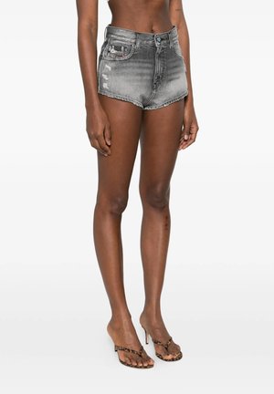 Persona che indossa shorts di jeans neri sbiaditi a vita alta e sandali con tacco alto a stampa leopardata in piedi su sfondo bianco.