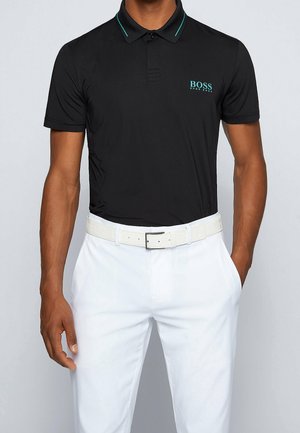 Mężczyzna w czarnej koszulce polo z krótkim rękawem z turkusowym logo i białych spodniach z białym paskiem, jedna ręka w kieszeni, na jednolitym tle.