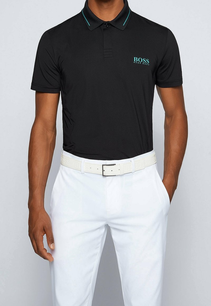 Homme portant un polo noir à manches courtes avec un logo turquoise et un pantalon blanc avec une ceinture blanche, une main dans la poche, sur un fond uni.