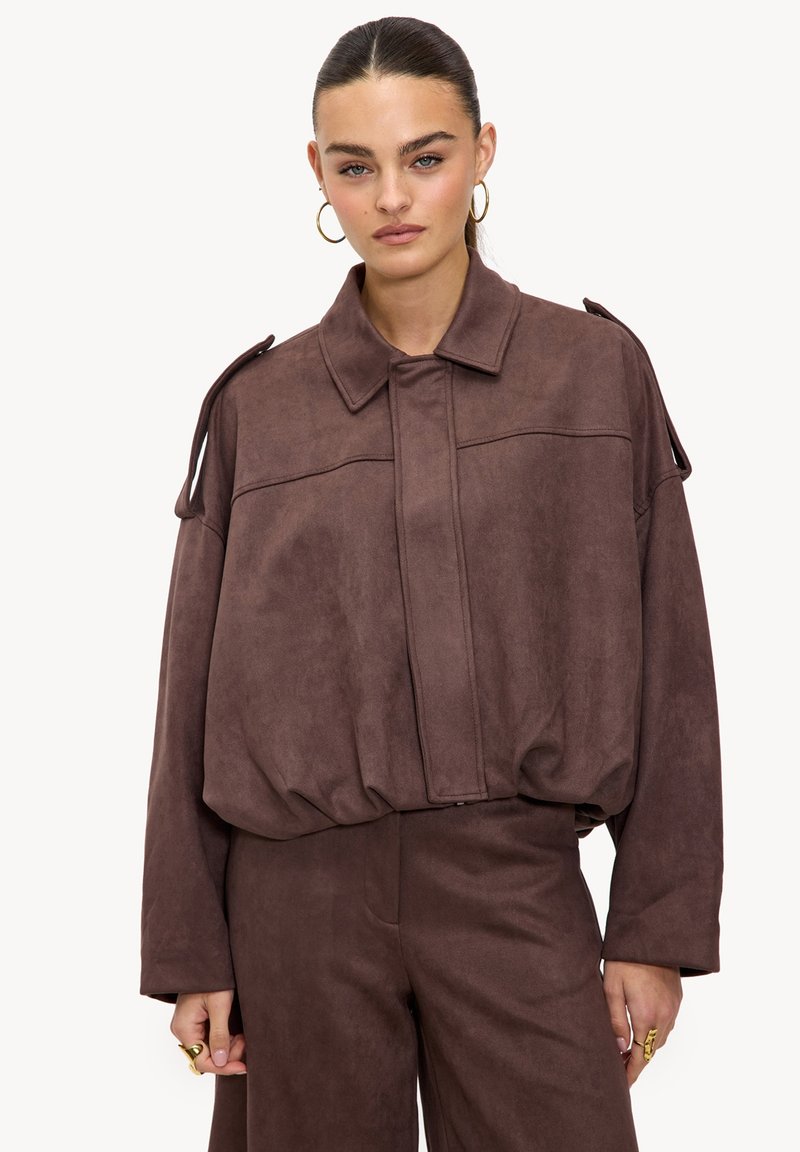 Veste marron en suédine synthétique avec une coupe décontractée, col arrondi et fermeture éclair. Dotée d'épaulettes aux épaules et de manches longues.