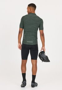 Kurzärmliges, dunkelgrünes Sportshirt mit einem strukturierten Design, kombiniert mit schwarzen Radshorts und einem schwarzen Helm in der Hand.