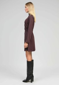 Femme blonde de profil, portant une robe longue à manches longues de couleur violet foncé et des bottes noires à talons hauts jusqu'aux genoux, sur fond gris clair.