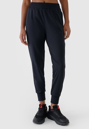 Osoba nosząca czarne sportowe spodnie dresowe typu jogger i czarne sneakersy z czerwonymi akcentami, stojąca na tle jednolitego jasnego tła.