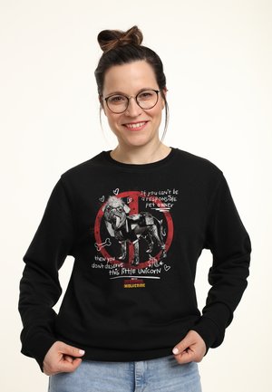 Donna sorridente con occhiali indossa una felpa nera con un'illustrazione di un cane e un testo sulla proprietà responsabile degli animali domestici.