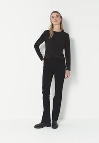 Schwarzes Langarmoberteil mit strukturierter Oberfläche, kombiniert mit figurschmeichelnden schwarzen Jeans und schwarzen Knöchelstiefeln, Modell steht vor einer weißen Wand.