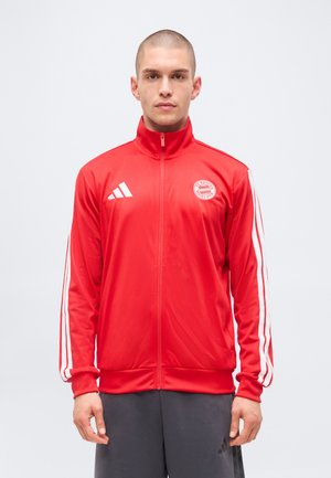 Hombre con chaqueta roja de FC Bayern Munich con cremallera, con logo blanco de Adidas y rayas, de pie frente a un fondo liso y claro.