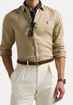 Mężczyzna w beżowej koszuli z guzikami, z podwiniętymi rękawami, granatową apaszką w kropki, białych plisowanych spodniach, brązowym pasku i zegarku na rękę.