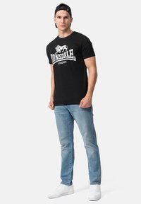 Sort bomuld t-shirt med et hvidt løve logo og teksten "LONSDALE LONDON", parret med lys blå jeans og hvide sneakers.