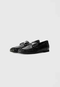 Sorte skinn loafers med rund tå, med sammenrullet detalj langs kanten og dekorasjoner foran. Flat såle.