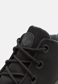 Zbliżenie na czarną tkaninę języka trampka z wyszywaną logo i czarnymi sznurówkami przewleczonymi przez metalowe oczka na jasnym tle.