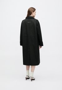 Palton negru lung dintr-un material neted, cu guler, umeri căzuți și o etichetă pe spate. Modelul poartă șosete deschise la culoare și pantofi cu toc.