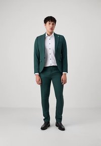 Costume vert avec une coupe ajustée, doté d'un revers cranté et d'une fermeture à un seul bouton, associé à une chemise blanche boutonnée et des chaussures noires.