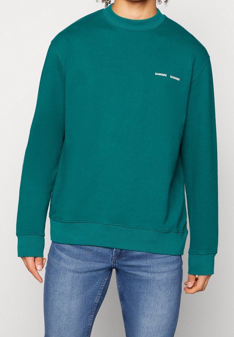 Modrý svetrový svetr z bavlněné směsi s žebrovanými manžetami a lemem. Malé vyšívané logo na hrudi. Doplňuje modré džíny.