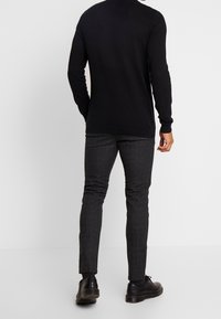 Suéter negro de manga larga con cuello redondo y dobladillo acanalado, combinado con pantalones taperados de cuadros gris oscuro y zapatos negros.