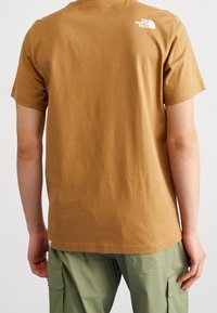 Brązowa bawełniana koszulka z okrągłym dekoltem i krótkimi rękawami, z białym logo na górnej części pleców, zestawiona z zielonymi spodniami cargo.