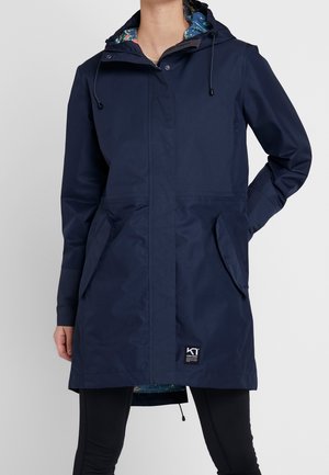 Femme portant un imperméable à capuche bleu marine avec poches avant et doublure fleurie, associé à des leggings noirs.