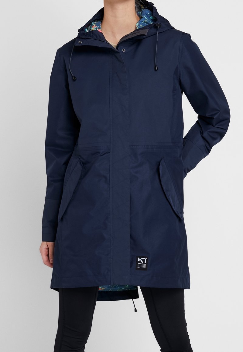Femme portant un imperméable à capuche bleu marine avec poches avant et doublure fleurie, associé à des leggings noirs.