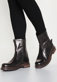 Botas de cuero negras hasta el tobillo con un acabado brillante, un zipper lateral y un acento de hebilla, que presentan una suela robusta y una entresuela marrón en contraste.
