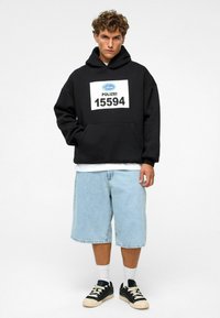 Haut à capuche noir avec une grande impression blanche "POLIZEI" et le numéro 15594, associé à un short en denim bleu clair de longueur genou et des baskets noires.
