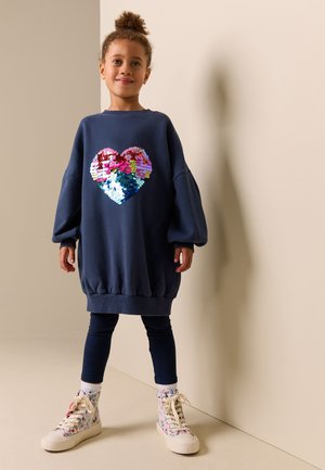 Jong meisje staat, draagt een oversized marineblauwe jurk met een meerkleurig paillettenhart, marineblauwe leggings en witte hoge sneakers met patronen.