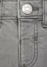 Țesătură de denim gri cu un buzunar vizibil, cusut cu fir întunecat și un buton metalic argintiu gravat cu "LAB MNG DENIM."