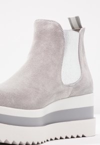 Harmaat mokkanahka nilkkurit, joissa on valkoinen joustava sivupaneeli, paksu valkoinen ja harmaa vaimennettu pohja sekä sahalaitatekstiili alareunassa.