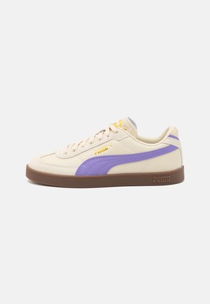 Baskets Puma couleur crème avec une bande violette, logo jaune, semelle marron et lacets sur fond blanc, vue latérale.