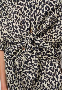 Leopard print lightweight blouse with a tie at the waist, featuring short sleeves and a button-down front. Color palette includes cream, black, and brown.