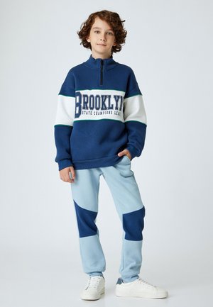 Sudadera azul con cremallera y rayas blancas y verdes, que presenta el texto "BROOKLYN". Pantalones de chándal azul claro con acentos en azul marino y textura acanalada. Zapatillas blancas.