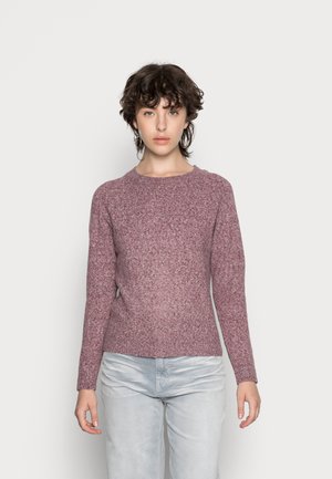 Person med kort, krøllet hår står vendt fremad iført en mauve striktrøje og lysegrå bukser mod en ensfarvet hvid baggrund.