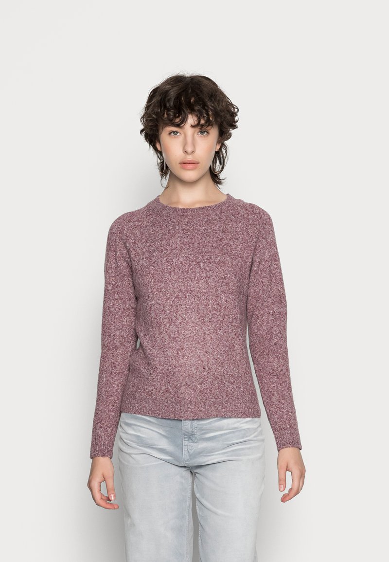 Personne aux cheveux courts et bouclés, debout face à l'avant, portant un pull en maille mauve et un pantalon gris clair, devant un fond blanc uni.
