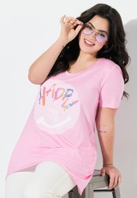 Camiseta rosa de manga corta con cuello redondo, que presenta un gráfico de lentejuelas multicolor con la inscripción "SOLO BUENAS VIBRACIONES". Combinada con pantalones blancos.
