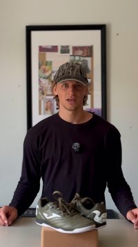 Mies, który nosi czapkę i czarną koszulę, stoi za parą oliwkowo-zielonych butów sportowych na brązowym pudełku, z tablicą ogłoszeniową w tle.